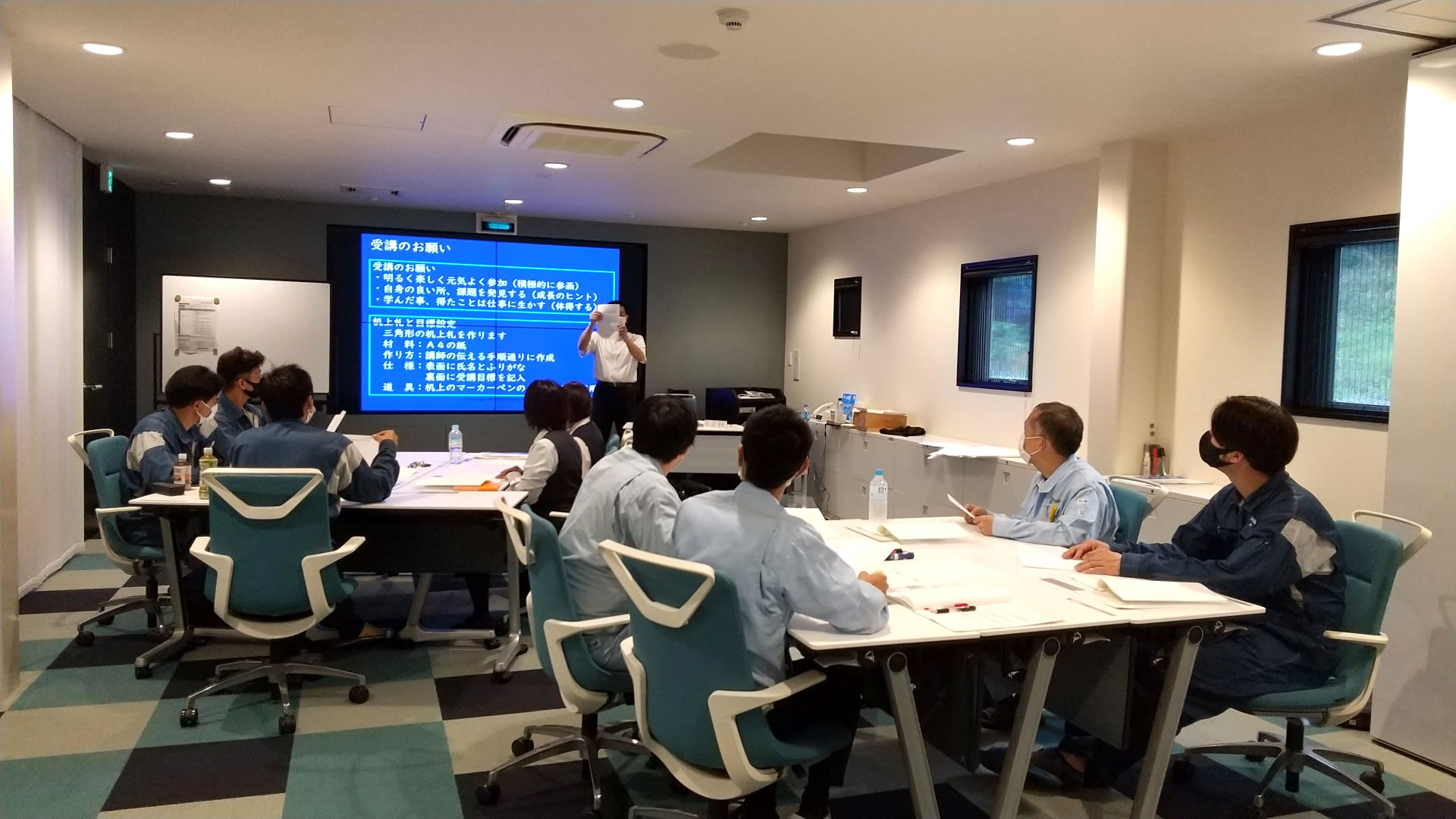 講習室に9名の社員が2つのテーブルに分かれて座っていて講師が前に立って電子黒板を使って救命講習をしている